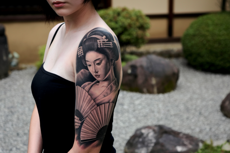 A photo of a Japanese woman with an ultra-realistic tattoo of a geisha. The tattoo covers her arm and shoulder, and is in black and grey. The geisha is holding a fan and has a serene expression. The background is blurred and consists of a traditional Japanese garden with rocks, gravel, and greenery.