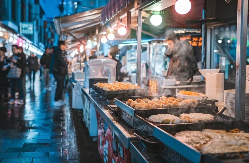 A photo of a lively Osaka street scene at night. The street is bustling with food stalls, and there are people in the background. The focus is on authentic Osaka street foods like takoyaki and okonomiyaki. There are sizzling street grills. Steam is rising from hot plates. The lights are colorful, and they reflect on the wet pavement. The atmosphere is energetic and urban.