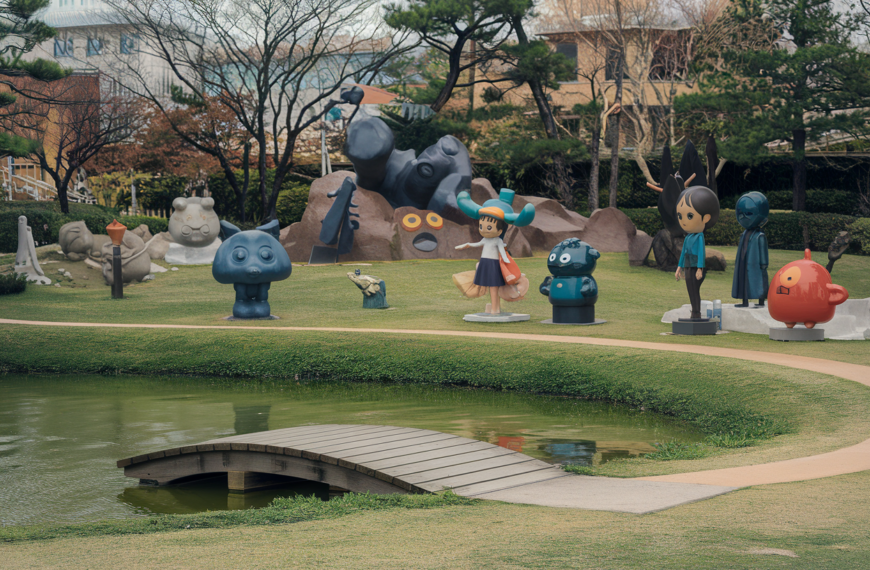 A photo of the Studio Ghibli Park in Japan. There are various sculptures of characters from the Studio Ghibli movies. The park has a small lake with a wooden bridge. The background contains trees and buildings. The ground is covered with green grass.