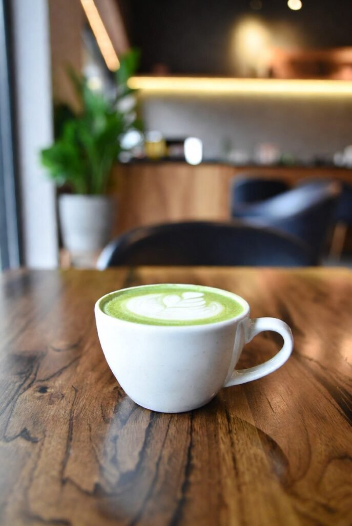 Matcha Kyoto: Exploring the Best Matcha 2026! 3 A photo of a cup of matcha latte in a modern
cafe. The latte is in a white cup with a dark green ring near the top. The cup is on a dark wooden surface. There is a small spoon next to the cup. The background has a few plants and a few more cups. The lighting is soft.