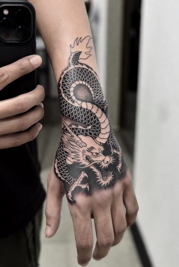 Person taking a mirror selfie showing a detailed traditional Japanese dragon tattoo across the hand, with bold scales and deep shading. The image is cropped to show only the hand, lit softly in a clean, minimal interior.