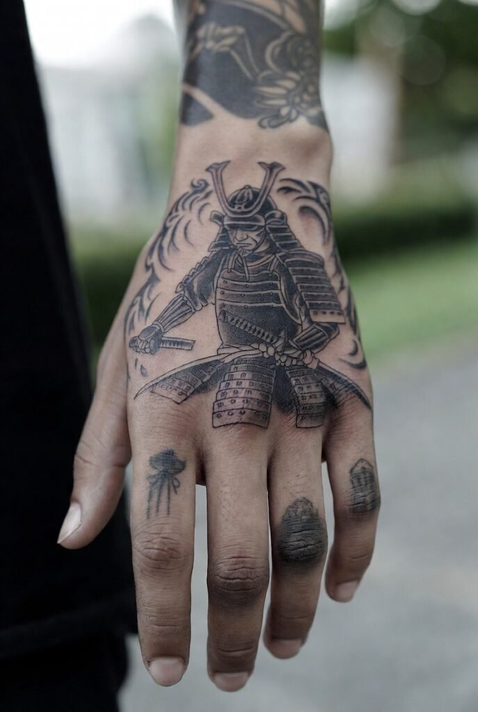 Person taking a mirror selfie highlighting a dynamic samurai figure holding a katana, positioned diagonally across the full hand. The face is not shown, with warm lighting emphasizing the bold line work.