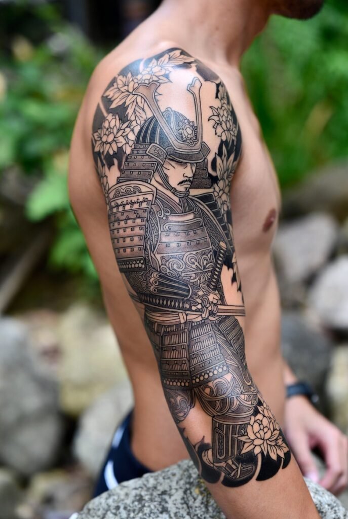 A Japanese model displays a detailed samurai tattoo on their arm in a blurred outdoor setting.