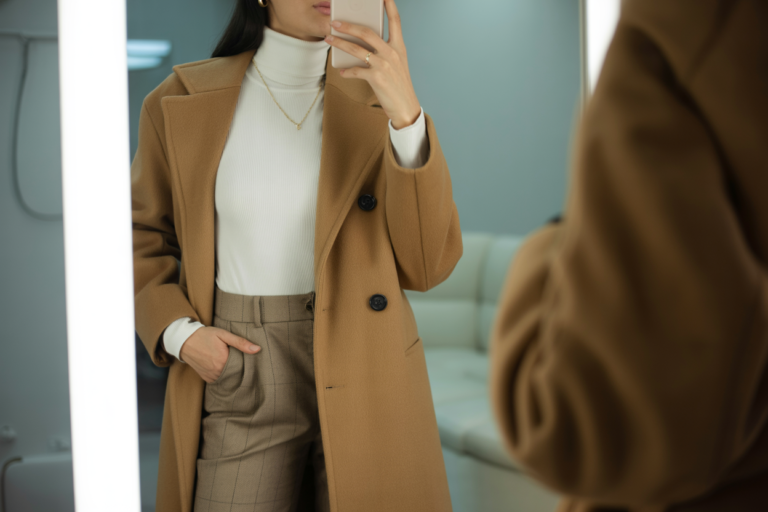 Japanese Winter Outfits Women: 10 Elegant Business Office Styles 2026! 12 Young woman taking a mirror selfie wearing a long camel wool coat over a fitted turtleneck and tailored trousers. Her face is cropped out, and the soft indoor lighting enhances the clean, executive winter atmosphere.