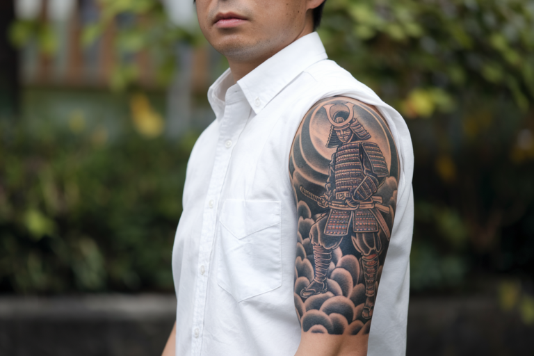 A medium shot of a Japanese man with a half sleeve tattoo of a Samurai on his arm. The man is wearing a white shirt and has short black hair. The tattoo is intricately detailed, with the Samurai holding a sword and standing on a cloud. The background is blurred and consists of greenery.