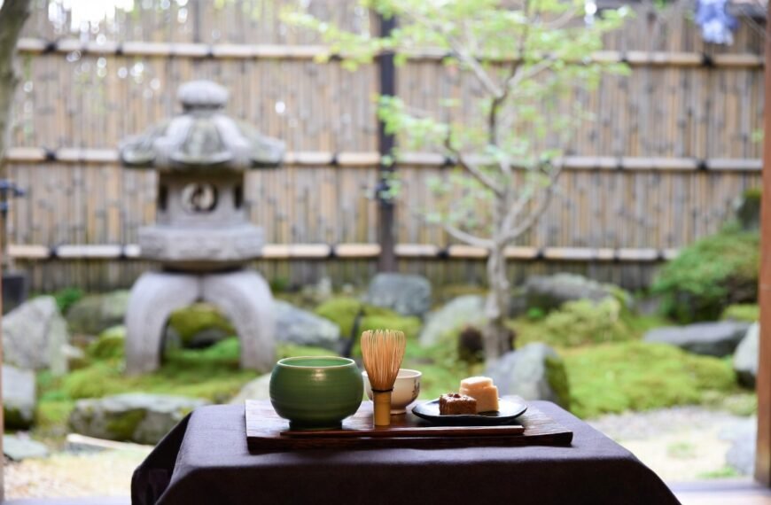 A photo of a cup of matcha latte in a modern cafe. The latte is in a white cup with a dark green ring near the top. The cup is on a dark wooden surface. There is a small spoon next to the cup. The background has a few plants and a few more cups. The lighting is soft.