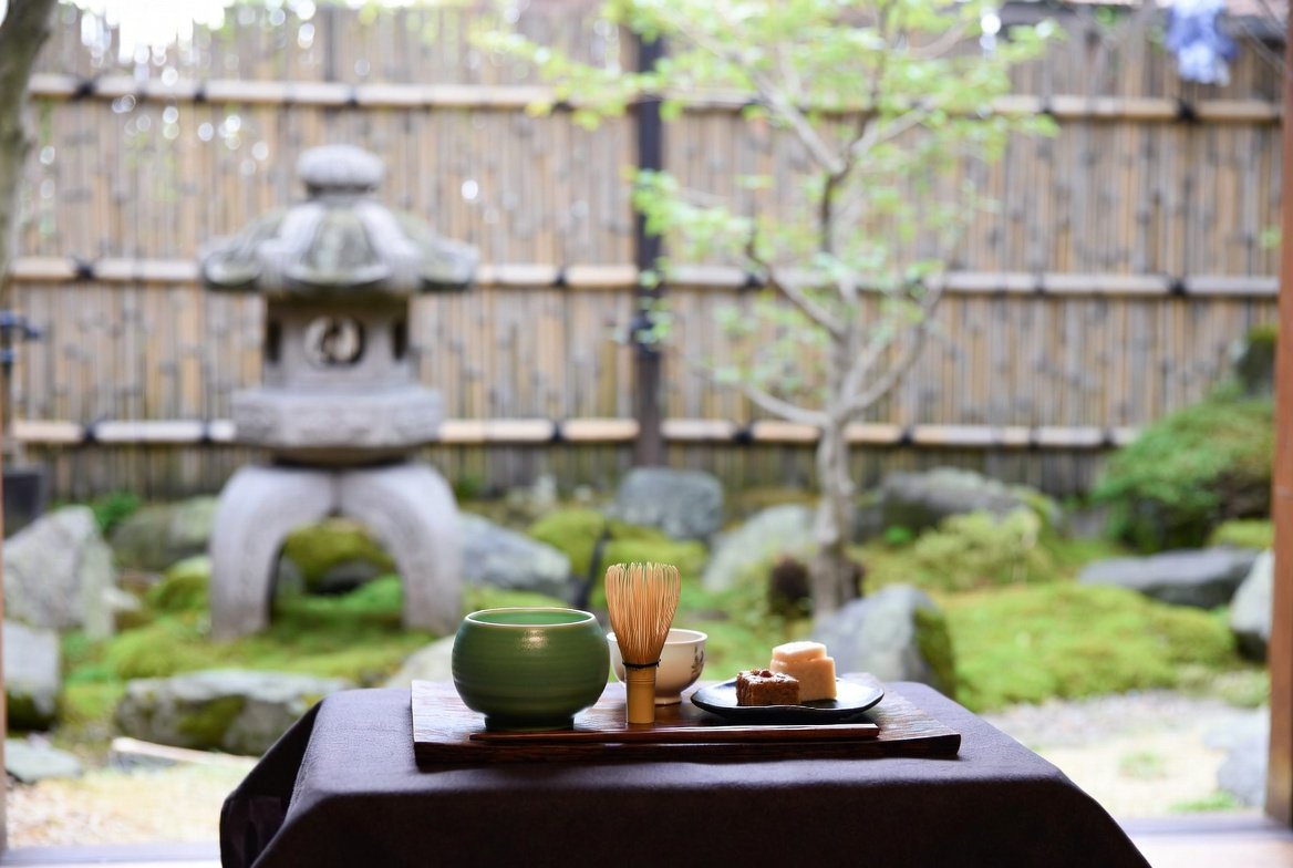 Matcha Kyoto: Exploring the Best Matcha 2026! 1 A photo of a cup of matcha latte in a modern cafe. The latte is in a white cup with a dark green ring near the top. The cup is on a dark wooden surface. There is a small spoon next to the cup. The background has a few plants and a few more cups. The lighting is soft.