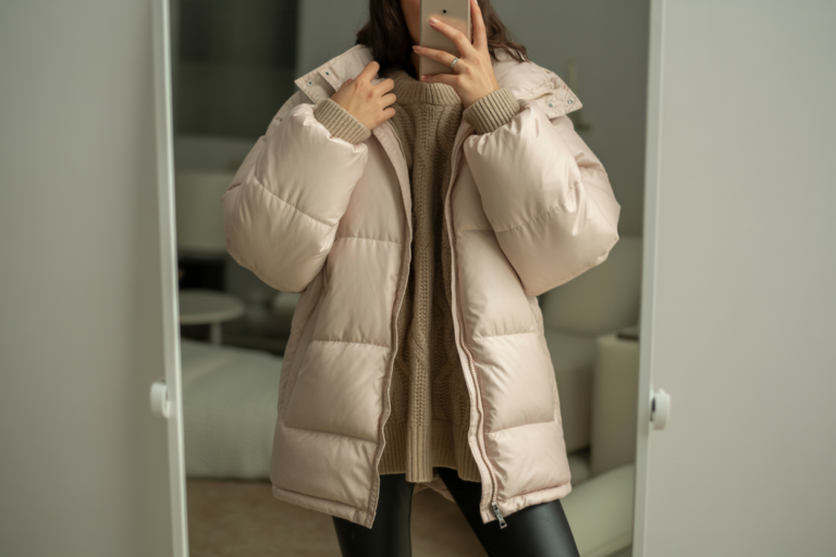 Winter Outfits Japanese: 10 Trendy Streetwear Styles for Young Women 2026! 13 Elegant young woman taking a mirror selfie wearing an oversized beige winter puffer with a cozy sweater and leggings. The photo shows her outfit without revealing her face, in soft indoor lighting with a clean, minimal background.
